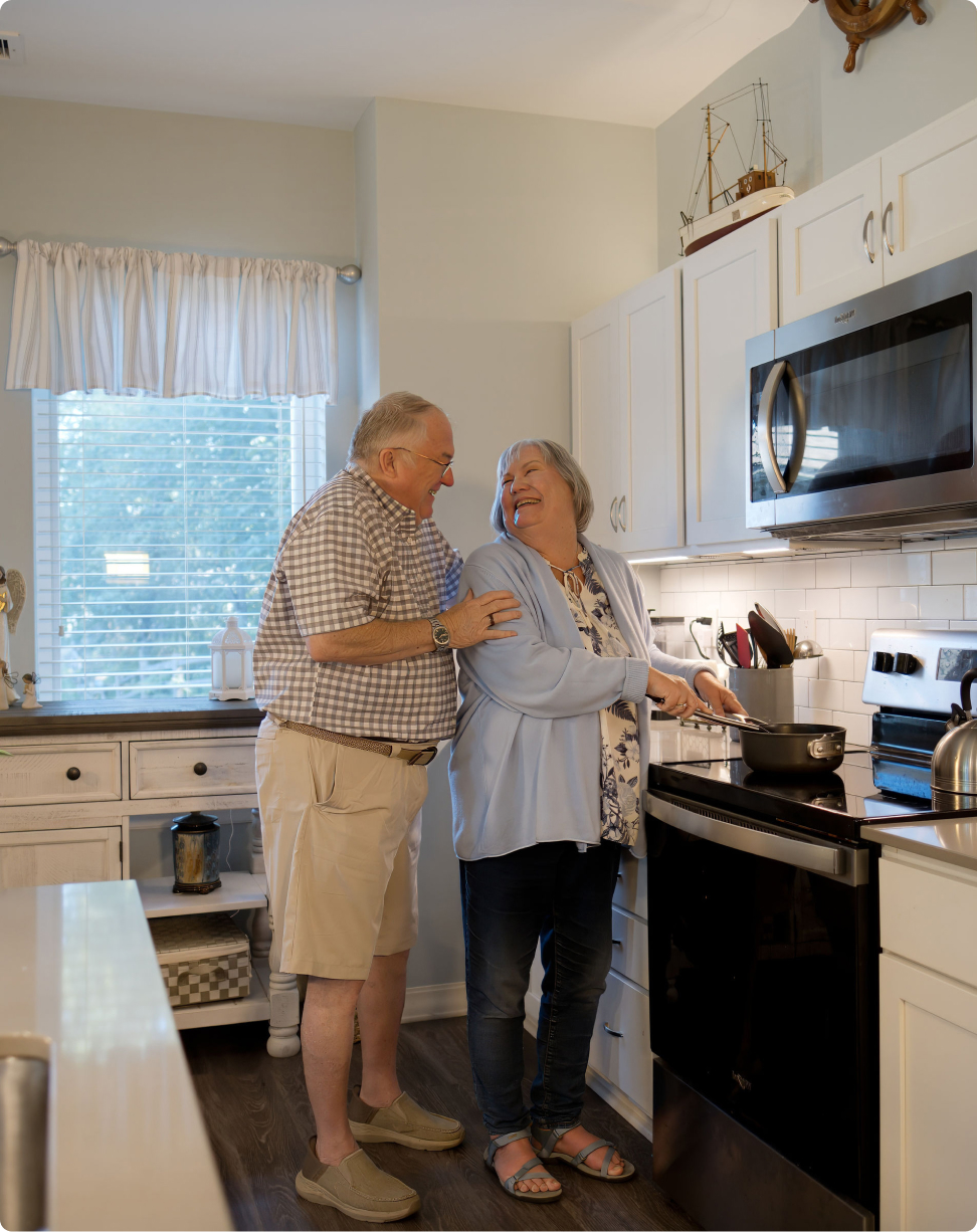 Two people smiling and cooking together in a cozy kitchen, featuring light cabinets and a window with natural light streaming in.