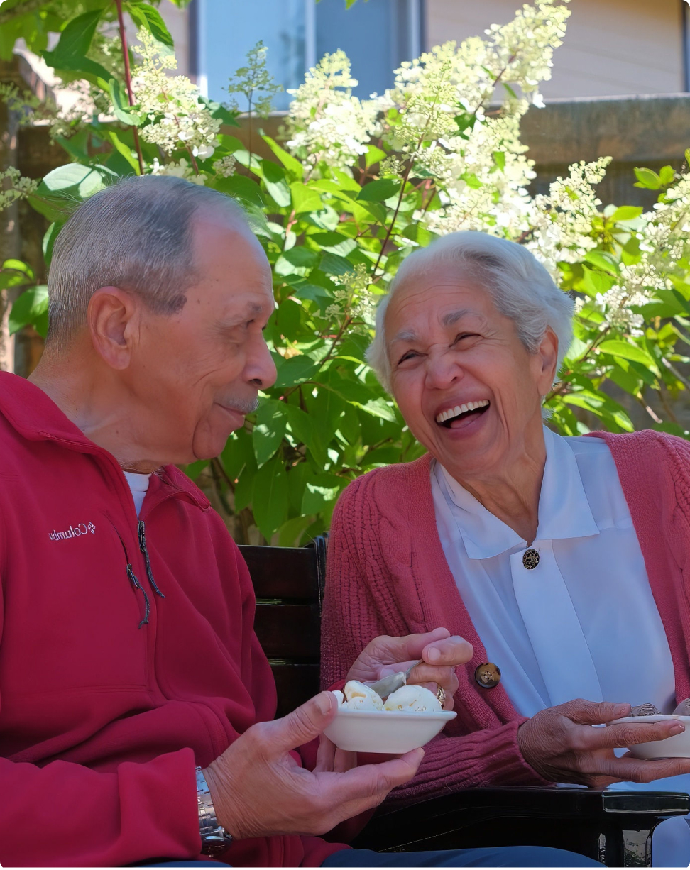 Two people laughing while enjoying ice cream on a bench, surrounded by vibrant green plants and white flowers in a sunny garden setting.