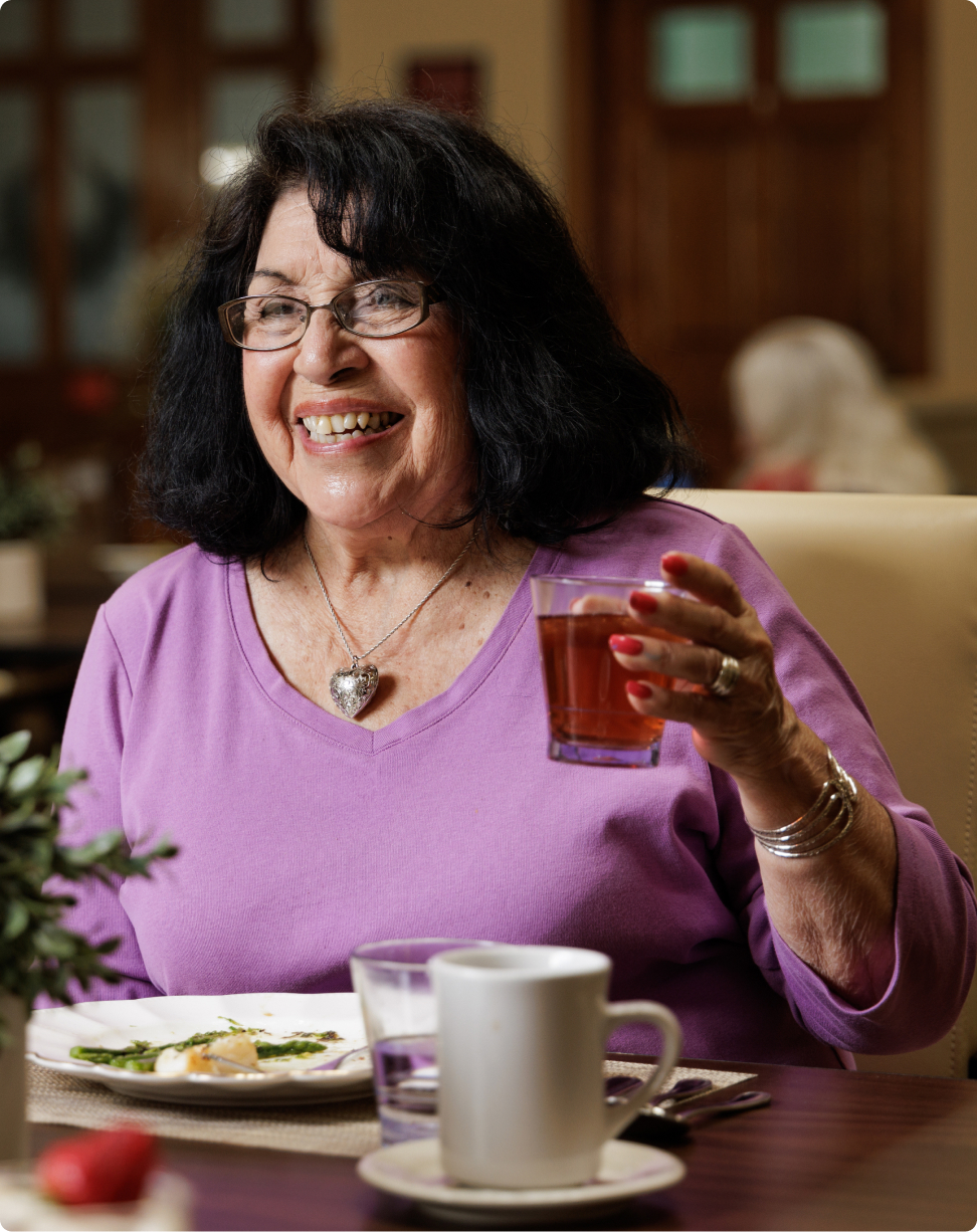 A person in a purple shirt smiles while holding a glass, seated at a dining table with plates and cups in a cozy room.