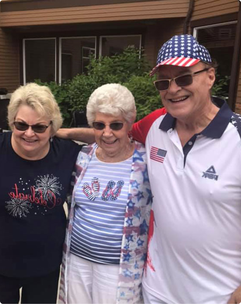 Three smiling people in patriotic attire, standing arm-in-arm outside a modern building with greenery. No recognizable landmarks or historical buildings visible.