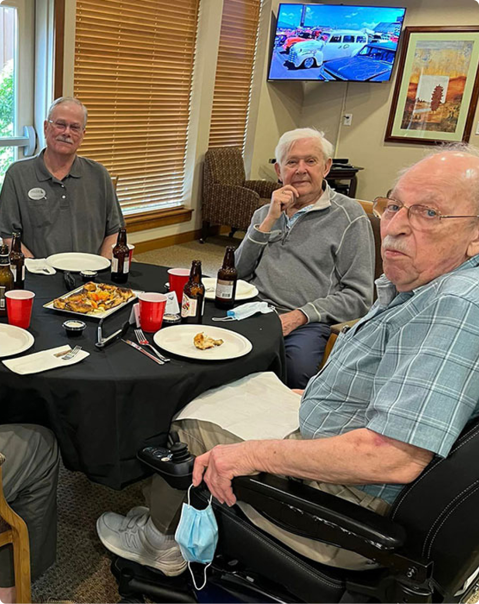 Three people gather around a table with pizza and drinks, watching a car show on TV in a cozy room.