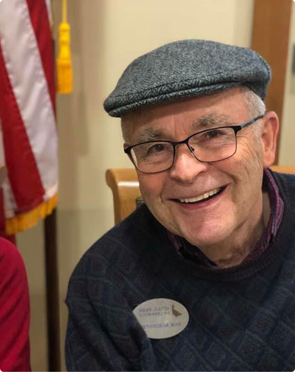 A smiling person wearing a cap and glasses, seated indoors near an American flag, wearing a name tag on their sweater.