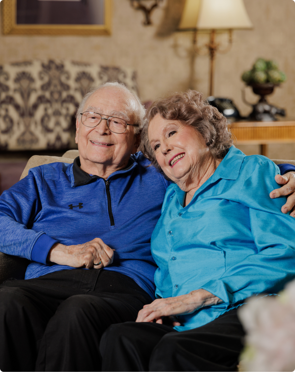 Two people sitting closely on a sofa, smiling warmly. An elegant room setting with patterned wallpaper and a vintage lamp in the background.