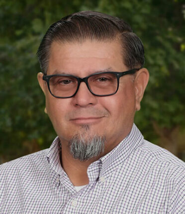 A person with glasses and facial hair is wearing a checkered shirt. The background features blurred greenery, suggesting an outdoor setting.