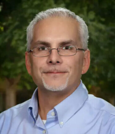 A person with glasses and short hair is wearing a light blue shirt, posing outdoors with greenery in the background.