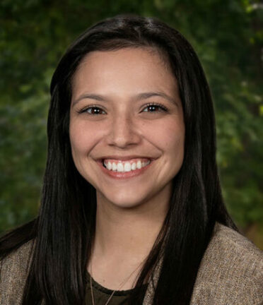 A person with long dark hair smiles against a blurred green background, wearing a brown jacket. No landmarks or historical buildings are visible.