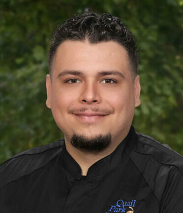 A person with short, curly hair and a goatee is wearing a black chef's jacket. The background shows green foliage.