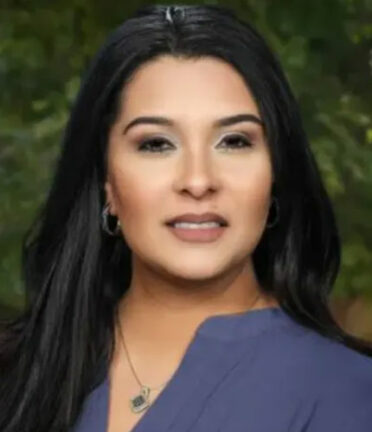 A person with long dark hair and a blue top is outdoors, with a blurred green background behind them.