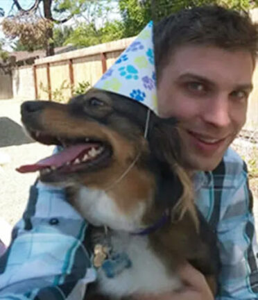 A person hugs a dog wearing a party hat in a backyard. Both are smiling. A wooden fence and trees are in the background.