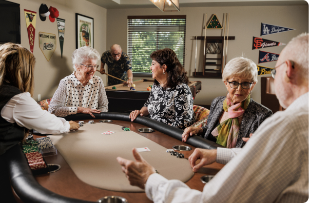 Five people gathered around a poker table with cards and chips, playfully engaged. Room decor includes sports pennants on the walls.