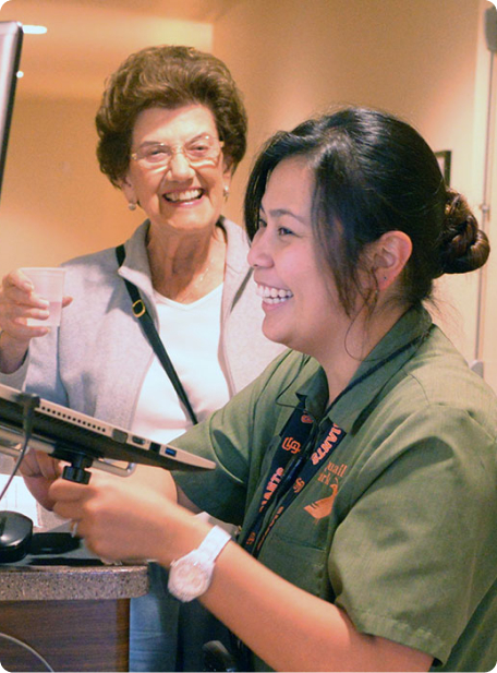 Two individuals smiling, one holding a tablet and the other with a cup. Indoor setting with warm lighting. No recognizable landmarks or historical buildings.