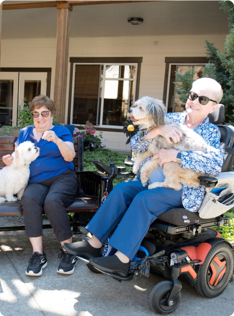 Two people with dogs sit outside on a sunny day, one in a wheelchair. Both appear happy and relaxed, enjoying the company of pets.