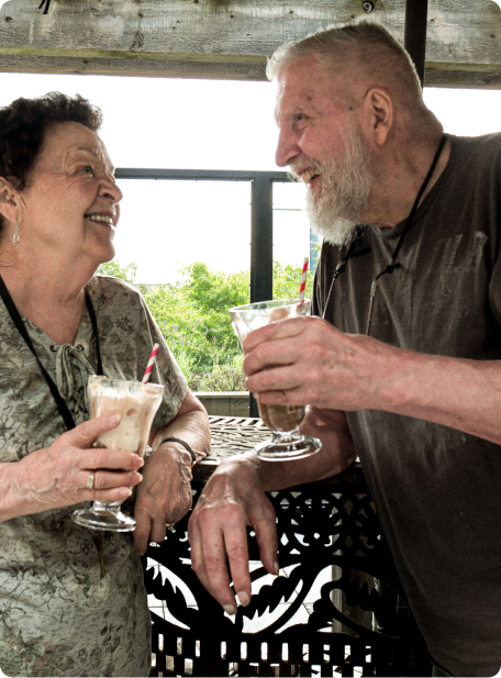 Two people smiling and holding milkshakes with straws, standing on a balcony with greenery in the background. Decorative table and railings visible.