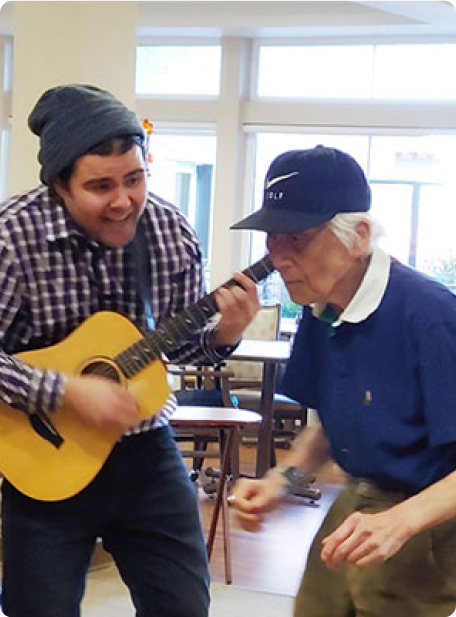 Two people are enjoying music indoors; one plays guitar, while the other dances. The scene suggests a lively, cheerful atmosphere.