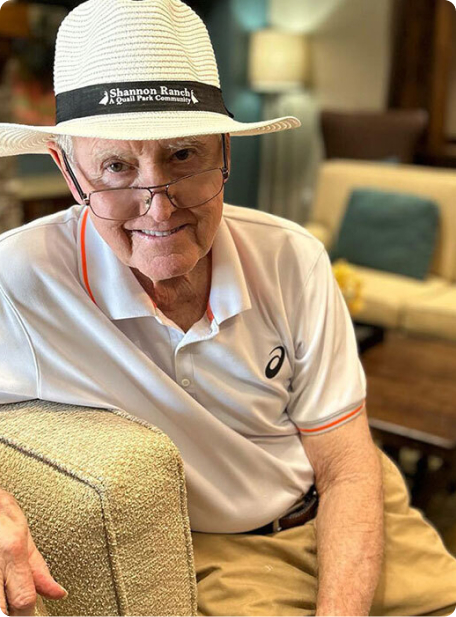 A person wearing a hat and glasses sits indoors on a couch, smiling. The room has soft lighting and cushioned furniture.