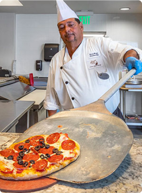 A person in a chef's uniform holds a pizza paddle, serving pepperoni pizza in a restaurant kitchen setting.