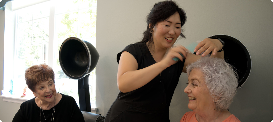 A person styling another person's hair in a salon, with two chairs and hairdryers. A third person is sitting nearby, smiling.