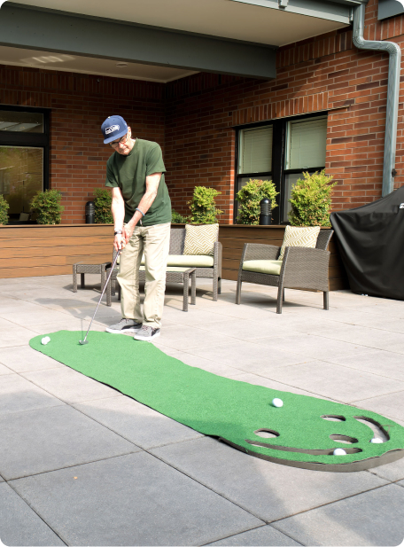 A person practices putting on a mini-golf mat in a brick patio courtyard, surrounded by potted plants and outdoor furniture.