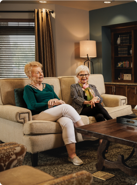 Two people are sitting on a sofa in a cozy, warmly lit living room with a lamp, bookshelves, and patterned decor.