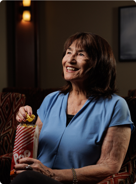 A person in a blue shirt smiles while holding a bag of popcorn, sitting indoors. The room has warm lighting and patterned furniture.