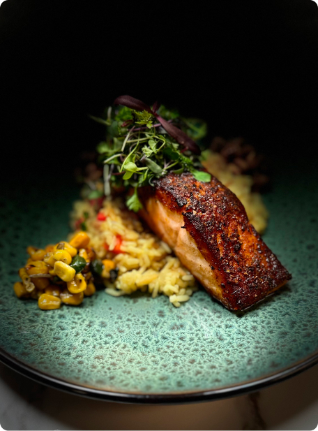 A perfectly seared salmon fillet is served on a textured green plate with creamy risotto, sautéed corn, and fresh herbs on top.