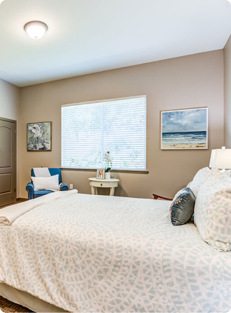 A cozy bedroom features a large bed, artwork, an armchair, and a window with blinds letting in natural light.