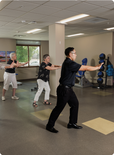 Three persons practice indoor exercises in a room with windows and equipment, including balance balls and chairs. They appear focused and synchronized.