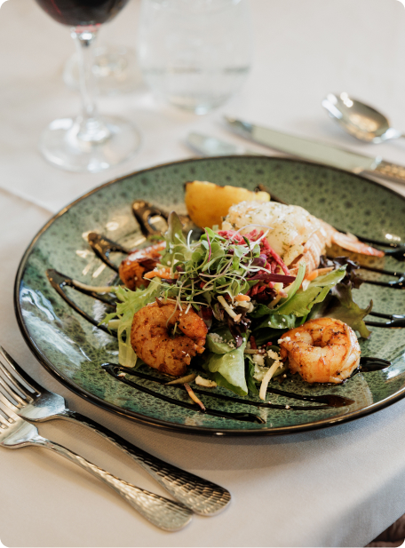 A culinary dish featuring grilled shrimp on a bed of greens, served with a glass of red wine on an elegant table setting.