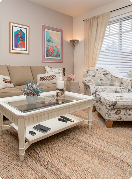 A cozy living room with beige sofa, patterned armchair, and glass-top coffee table. Decor includes framed artworks, lamps, and neutral curtains.