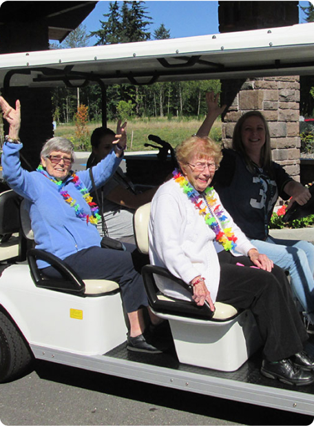 Three people wearing colorful leis ride in a golf cart, smiling and waving. There's greenery and a stone structure in the background.