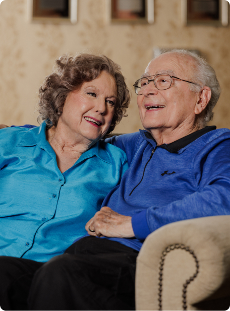 Two people sitting together on a couch, sharing a happy moment. Both are smiling, wearing casual blue clothing, inside a cozy room.