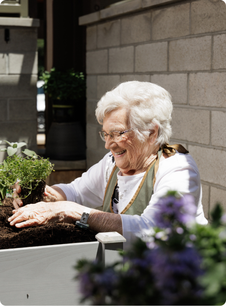 A person with white hair happily tends to plants in a garden. Stone wall and blurred greenery visible in the background.