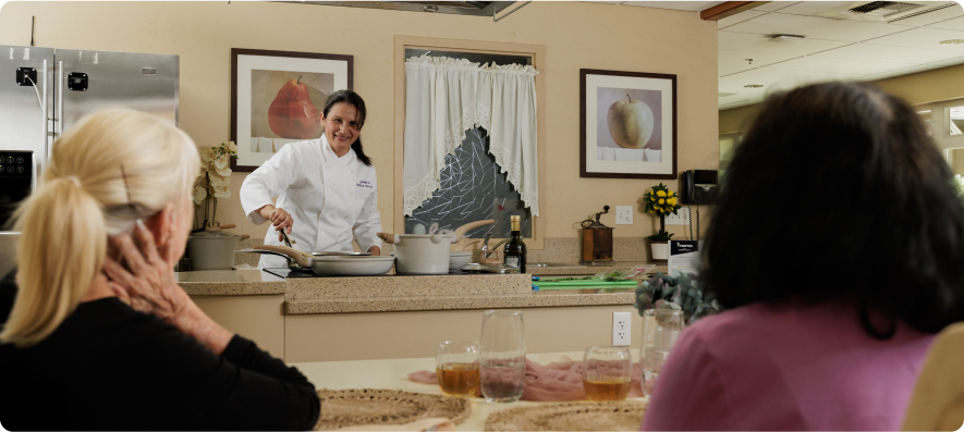 A person in a chef's coat demonstrates cooking in a kitchen while two people watch. Paintings and kitchen appliances are visible.