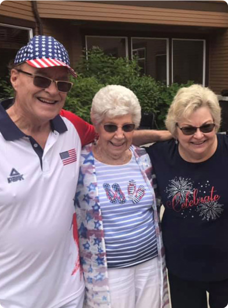 Three people are smiling outdoors, wearing festive clothing with American flag designs. They stand close together, with greenery and a house in the background.