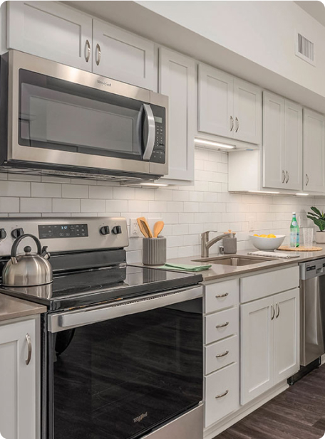 Modern kitchen with stainless steel appliances, white cabinetry, and subway tile backsplash. Includes microwave, stove, dishwasher, and countertop accessories like a kettle and bowl.