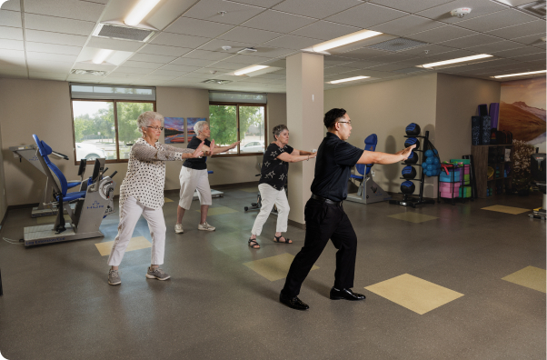 Four people practice tai chi in a fitness room with exercise equipment and scenic artwork on the walls.