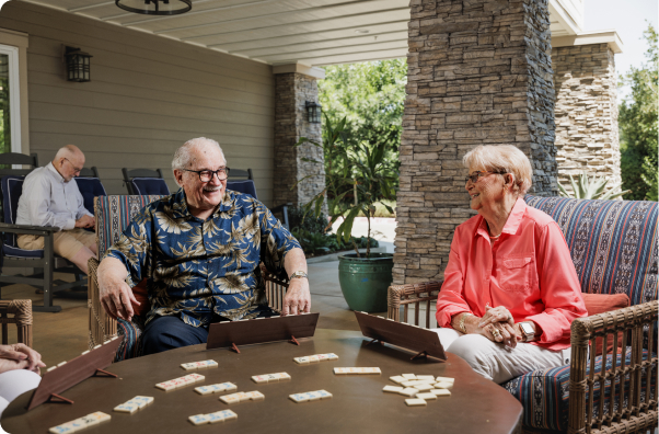 Two people sit outdoors on a patio, smiling and playing a tabletop game. A third person in the background is seated reading.