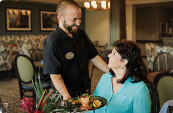 A person with a nametag serves a meal to another person in a warmly lit restaurant with patterned chairs and wall art.