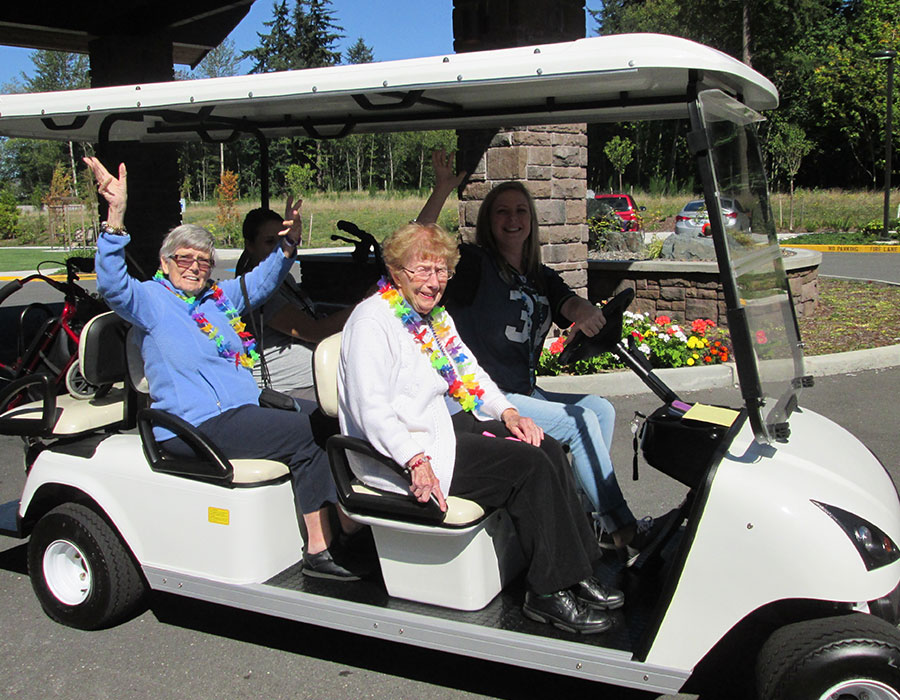 Three people wearing colorful leis enjoy a ride in a golf cart, surrounded by greenery and flowers in a sunny outdoor setting.