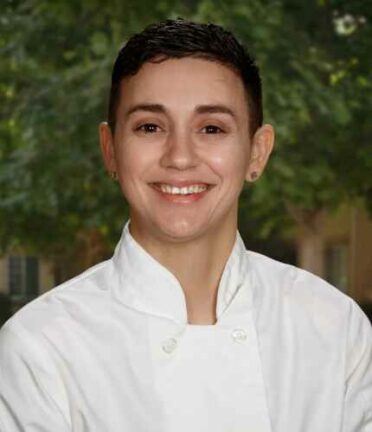 A person wearing a white chef's coat smiles against a blurred green, leafy background, likely outdoors. No recognizable landmarks or historical buildings visible.