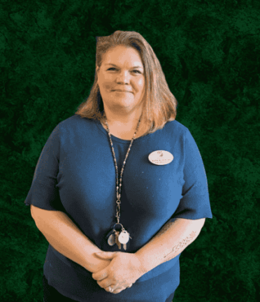 A person with shoulder-length hair wears a blue shirt and badge, standing against a dark green background, holding keys and a lanyard.