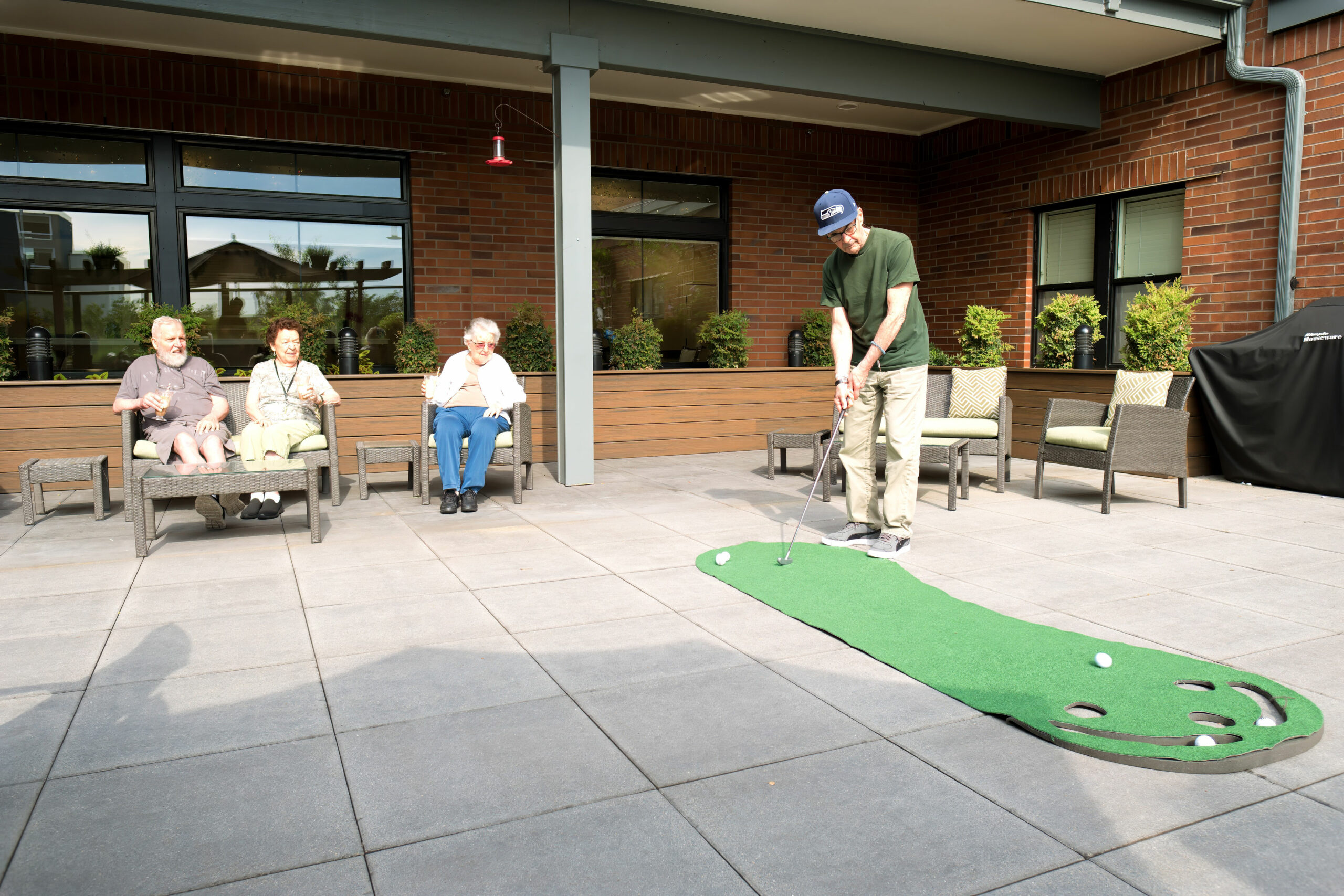 Four people relax on a patio; one plays mini-golf while three watch and enjoy seating at a retirement community or care facility.