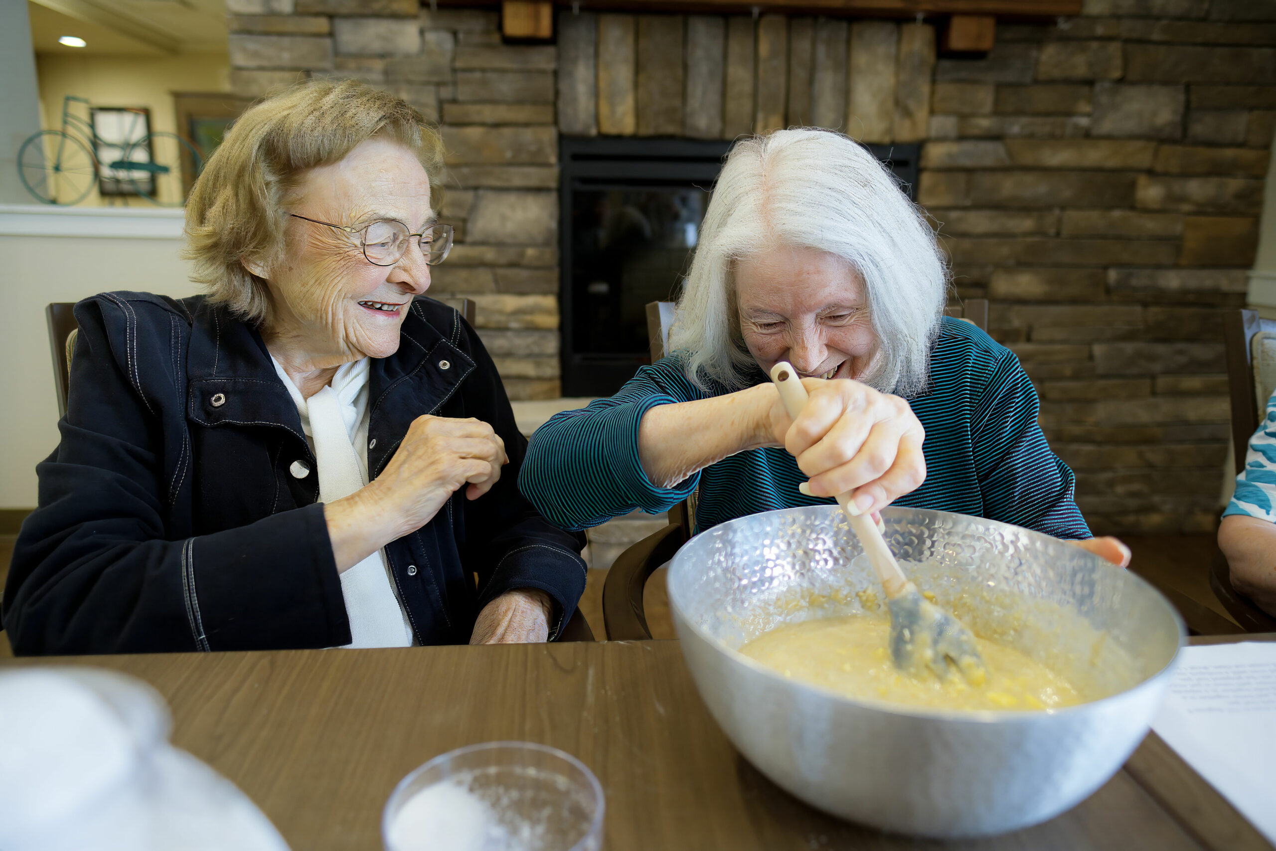 Two people joyfully mix ingredients in a large bowl at a wooden table, with a cozy stone fireplace in the background.