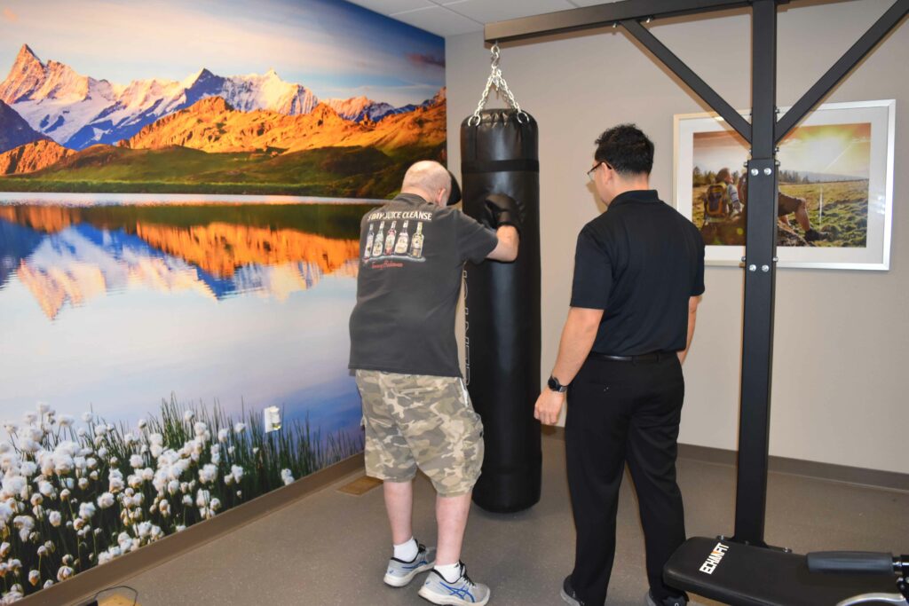 Two people near a punching bag; one person hits while the other observes. The room has a mountain mural and scenic photograph.